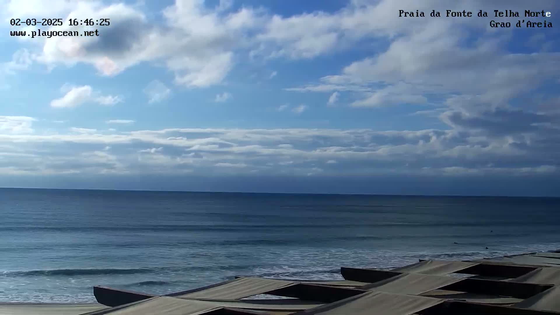 Under a partly cloudy sky, a calm ocean meets a beach where several covered structures are lined up.
