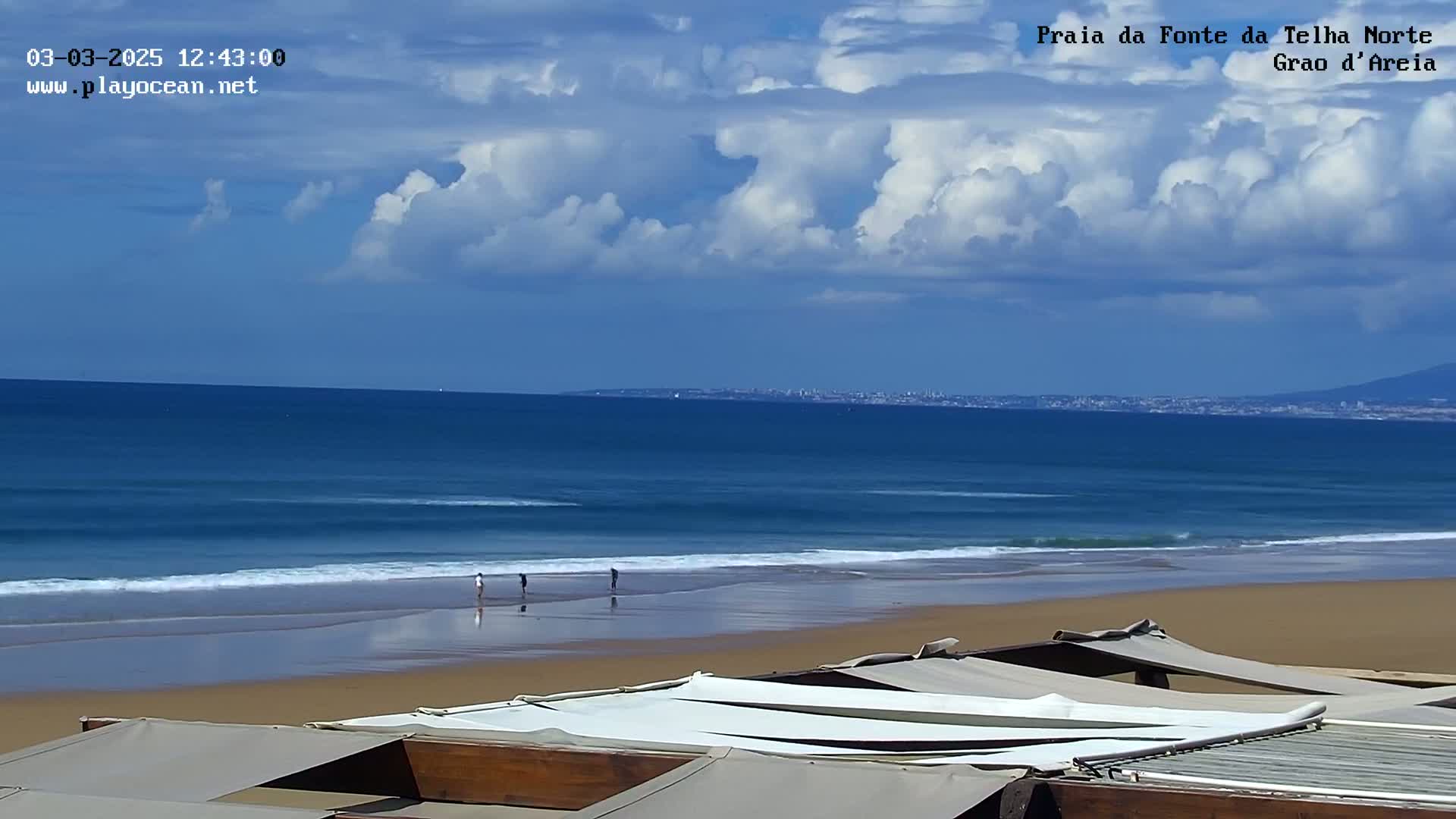 A sandy beach with a few people walking along the shore, under a partly cloudy sky, overlooks a blue ocean with gentle waves and a distant cityscape.