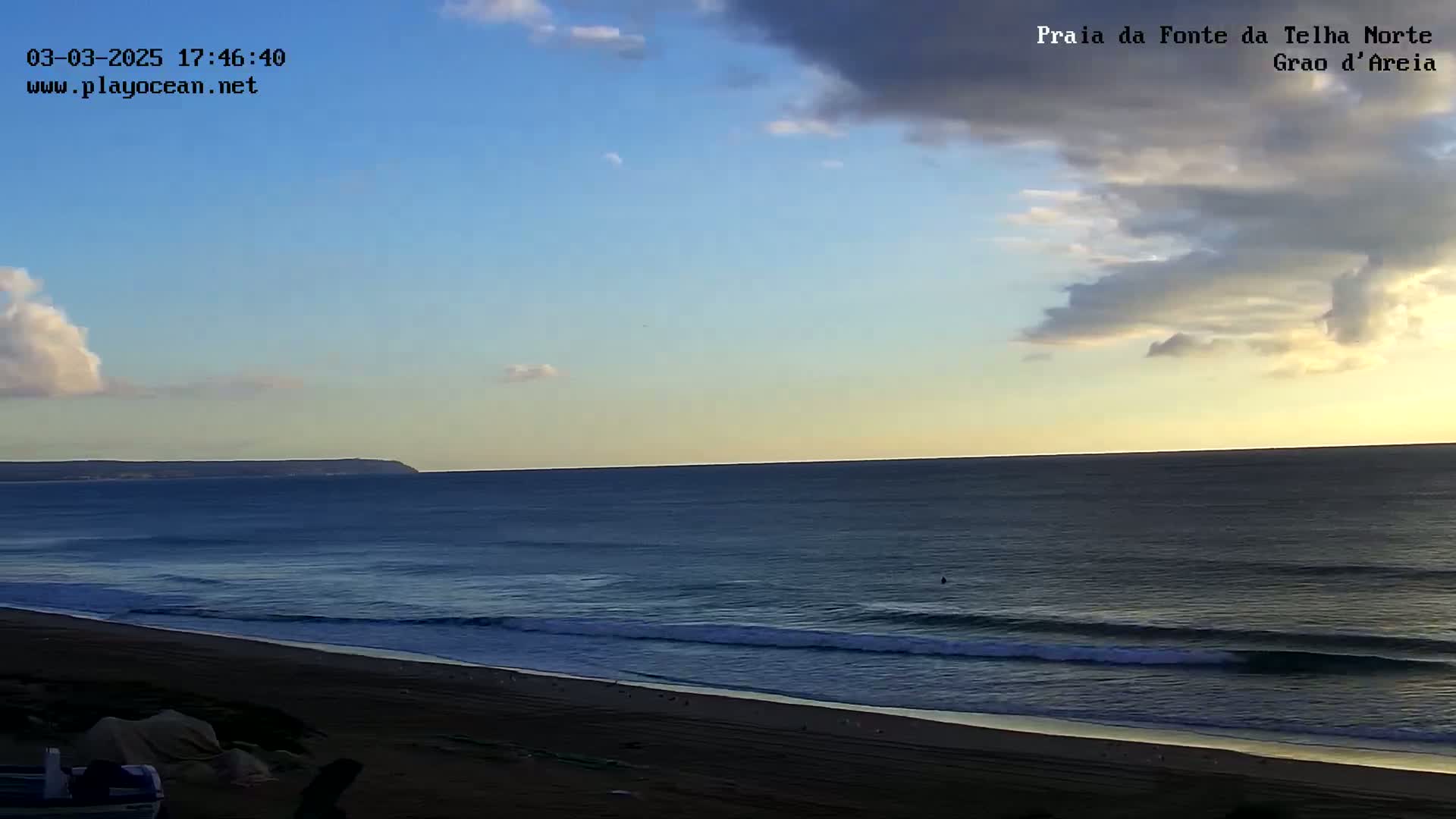 A partly cloudy sunset illuminates a wide sandy beach and ocean with gentle waves rolling onto the shore.