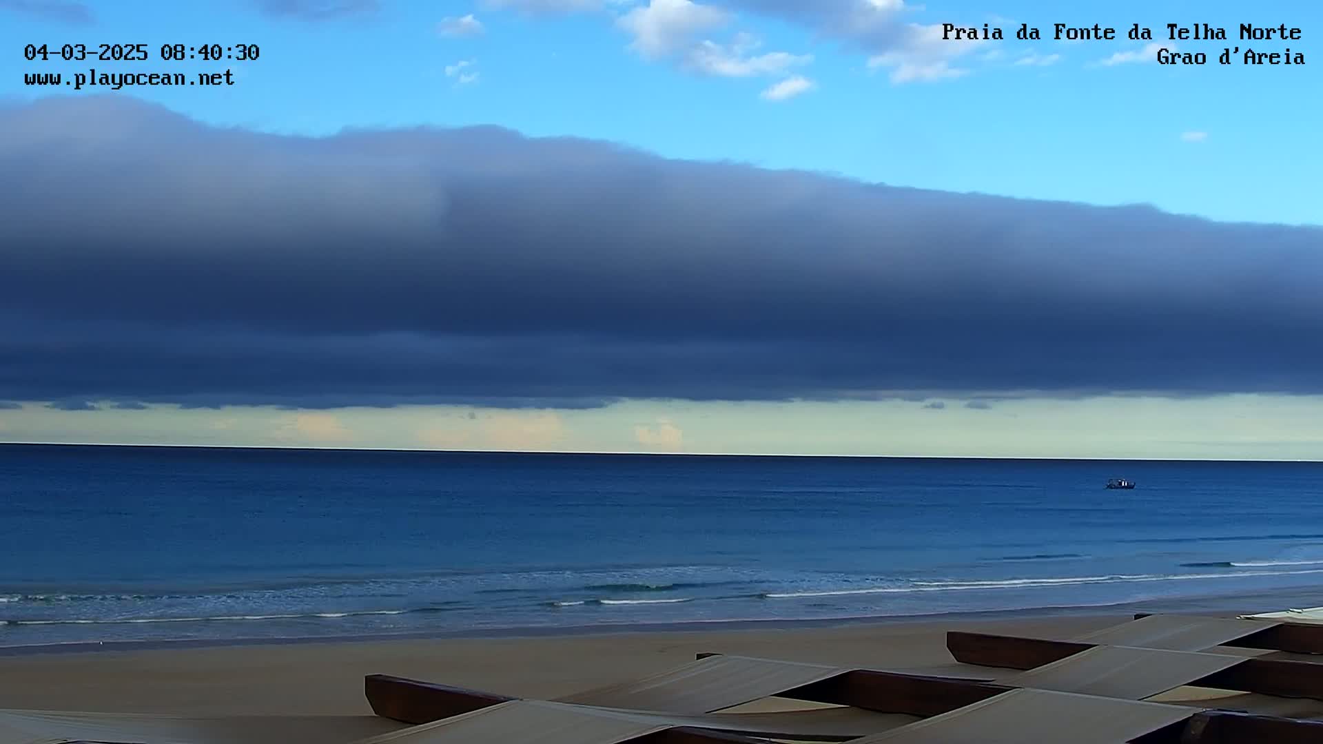 A dark, flat cloud layer hangs over a calm, blue ocean, with a sandy beach and several lounge chairs in the foreground.
