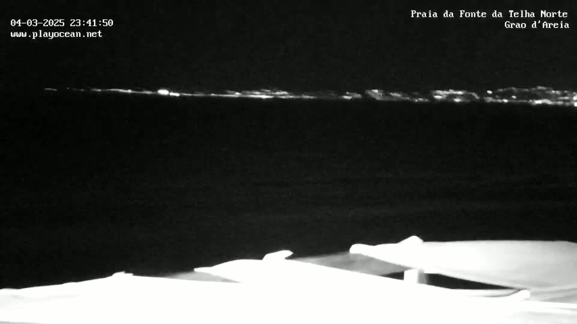 At night, a dark body of water shows a faint line of lights on the horizon, and in the foreground are two bright, indistinct objects.