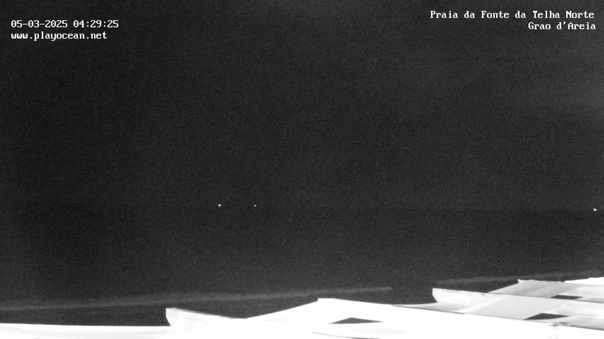 A nighttime, low-light view of a dark ocean with a few faint lights visible in the distance and the tops of white structures in the foreground.
