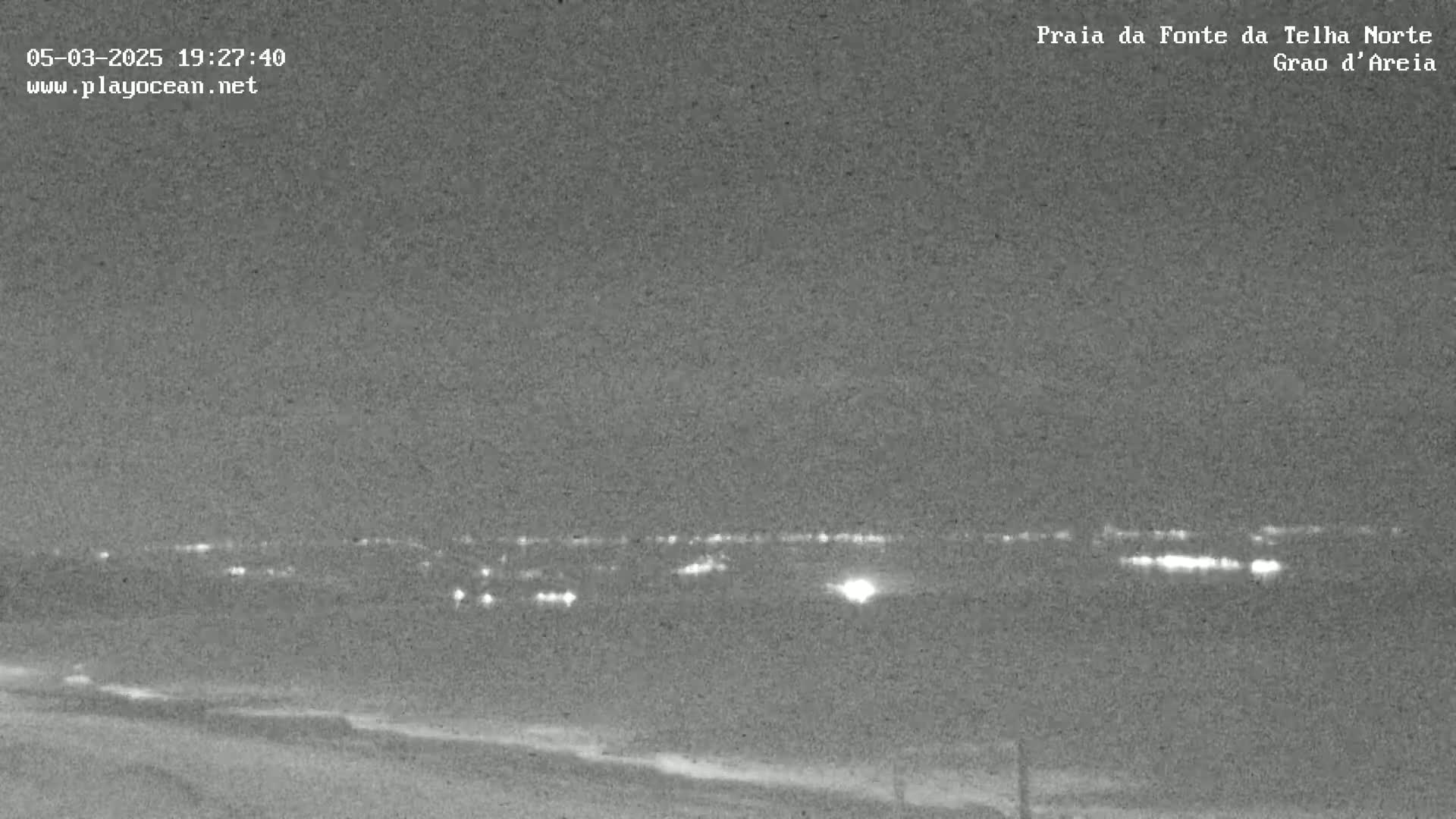 A nighttime, low-light image shows a dark beach and a distant line of lights in the background.