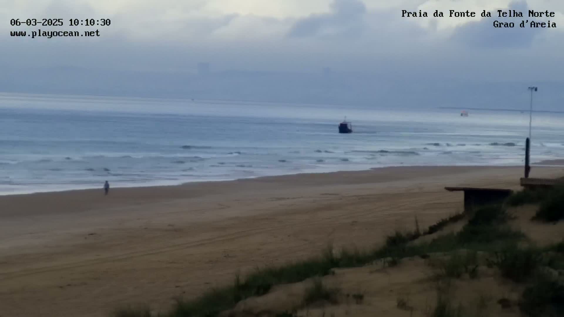 A lone person walks along a sandy beach with gentle waves rolling in under an overcast sky, a small vessel visible in the distance.