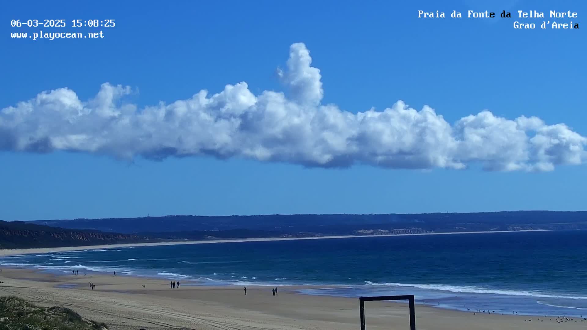 A sandy beach with gently rolling waves stretches beneath a mostly clear blue sky with a long, fluffy cloud formation.