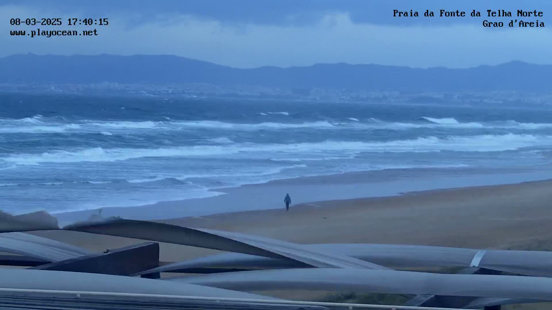 A lone figure walks on a wide, sandy beach under a cloudy sky, with a choppy ocean extending to distant mountains.