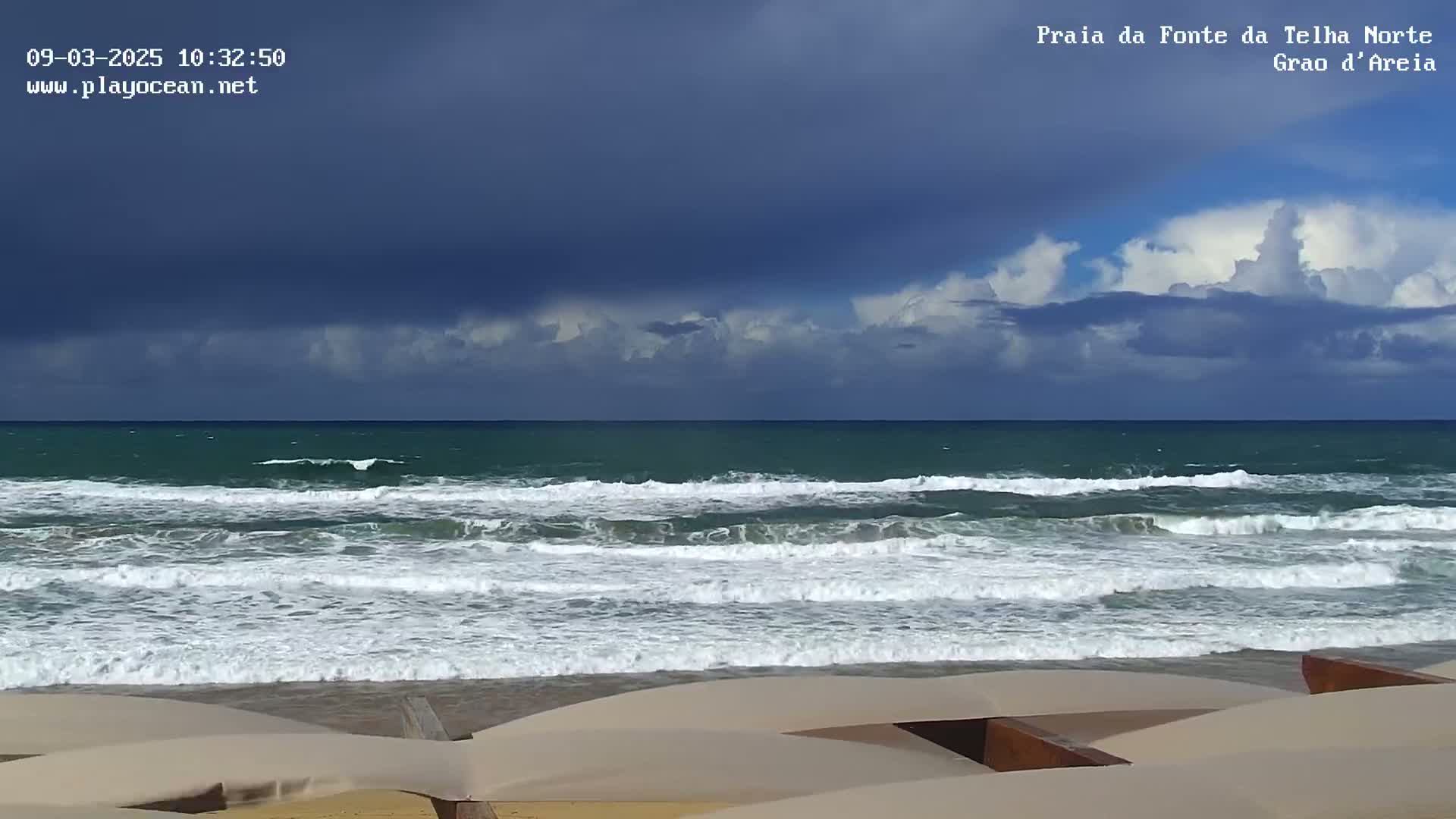 A beach scene shows dark, heavy clouds over a teal ocean with moderate waves breaking on a sandy shore where tan beach umbrellas are partially visible.