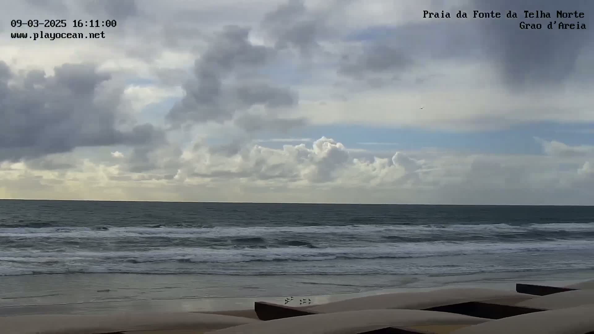 A mostly cloudy sky overlooks a wavy ocean meeting a sandy beach with several dark-colored objects resting on the sand.