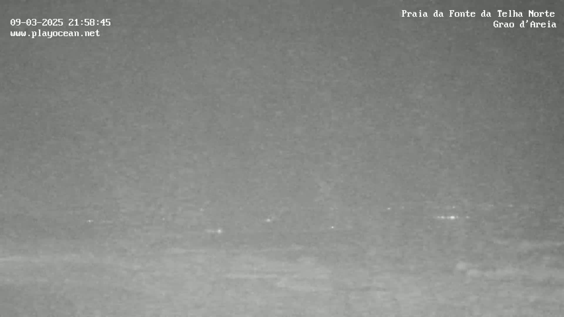 A nighttime, low-light image shows a dark, flat expanse of land or water, with a few faint lights visible in the distance.