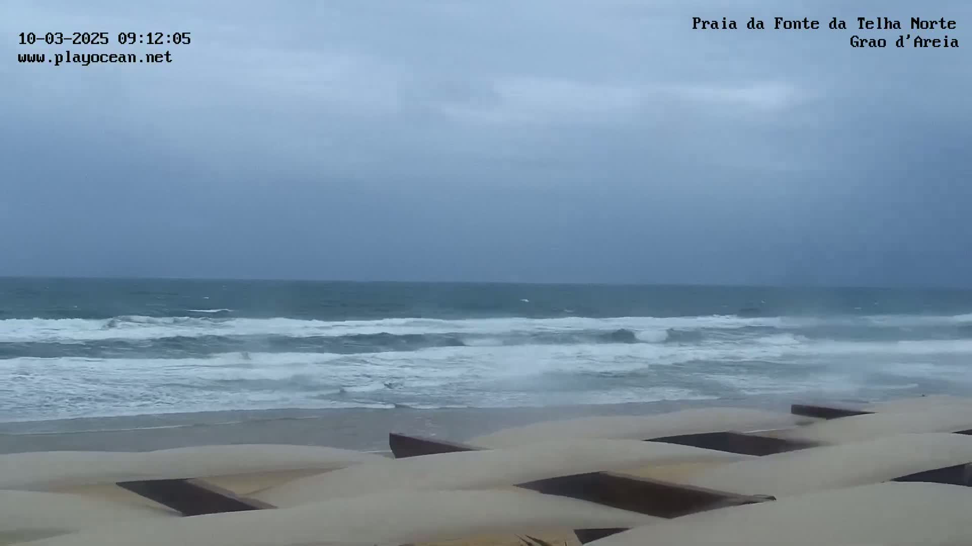 A beach with moderately rough waves under an overcast sky is partially obscured by a sandy dune structure in the foreground.