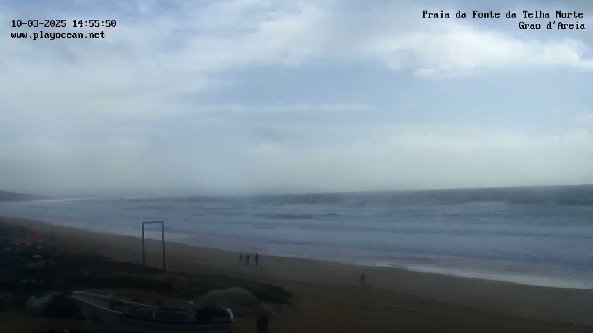A hazy, overcast day at a beach shows a mostly calm ocean with small waves, several people walking on the sand, and some vehicles parked near the shore.