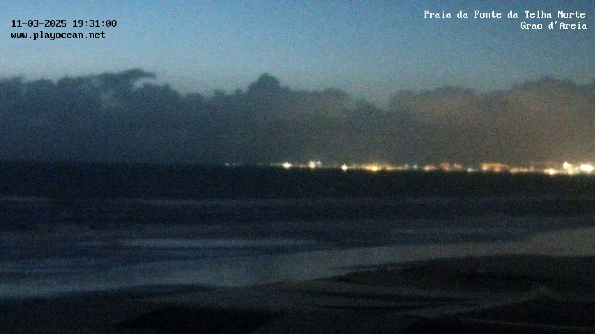 A dark, nighttime beach scene shows a dark ocean with a distant, faintly lit shoreline under a cloudy sky.