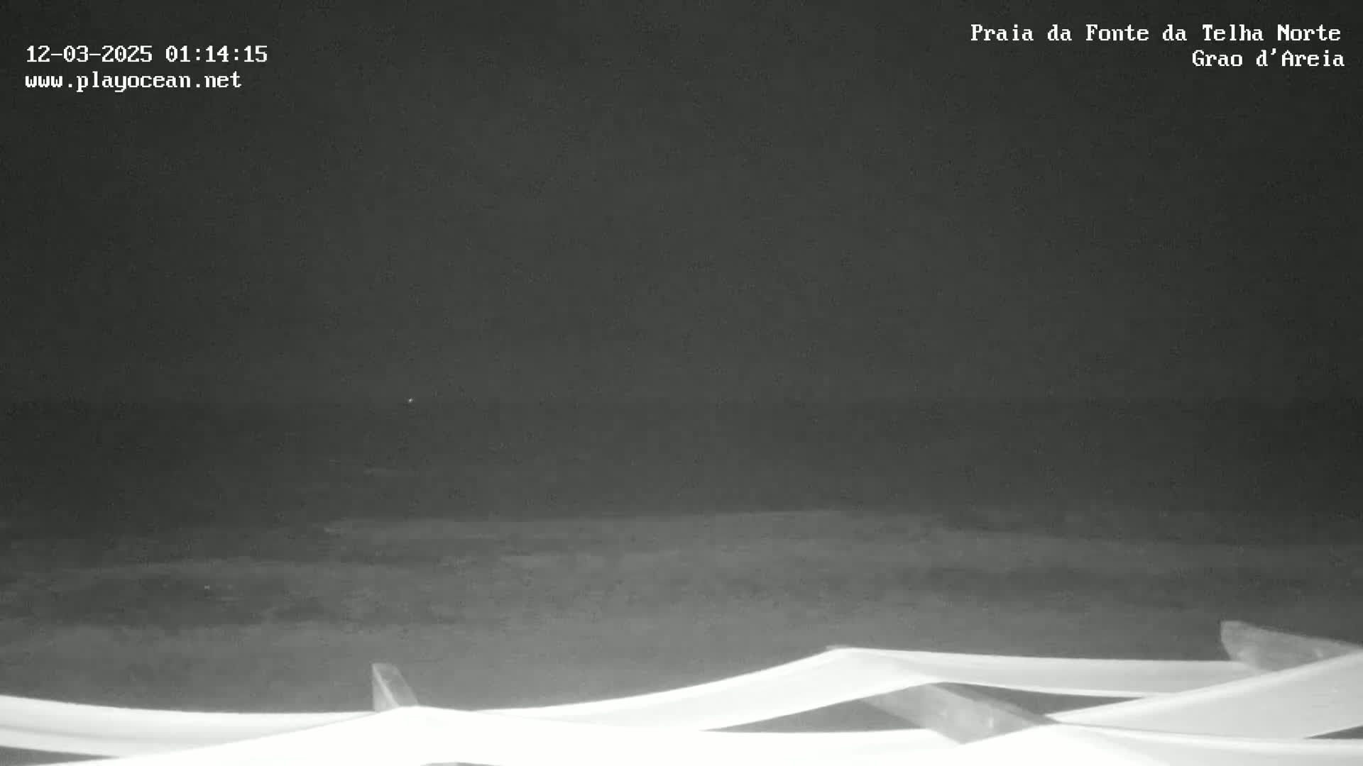 A dark, nighttime beach scene shows a mostly calm body of water under a black sky, with a section of white fabric in the foreground.