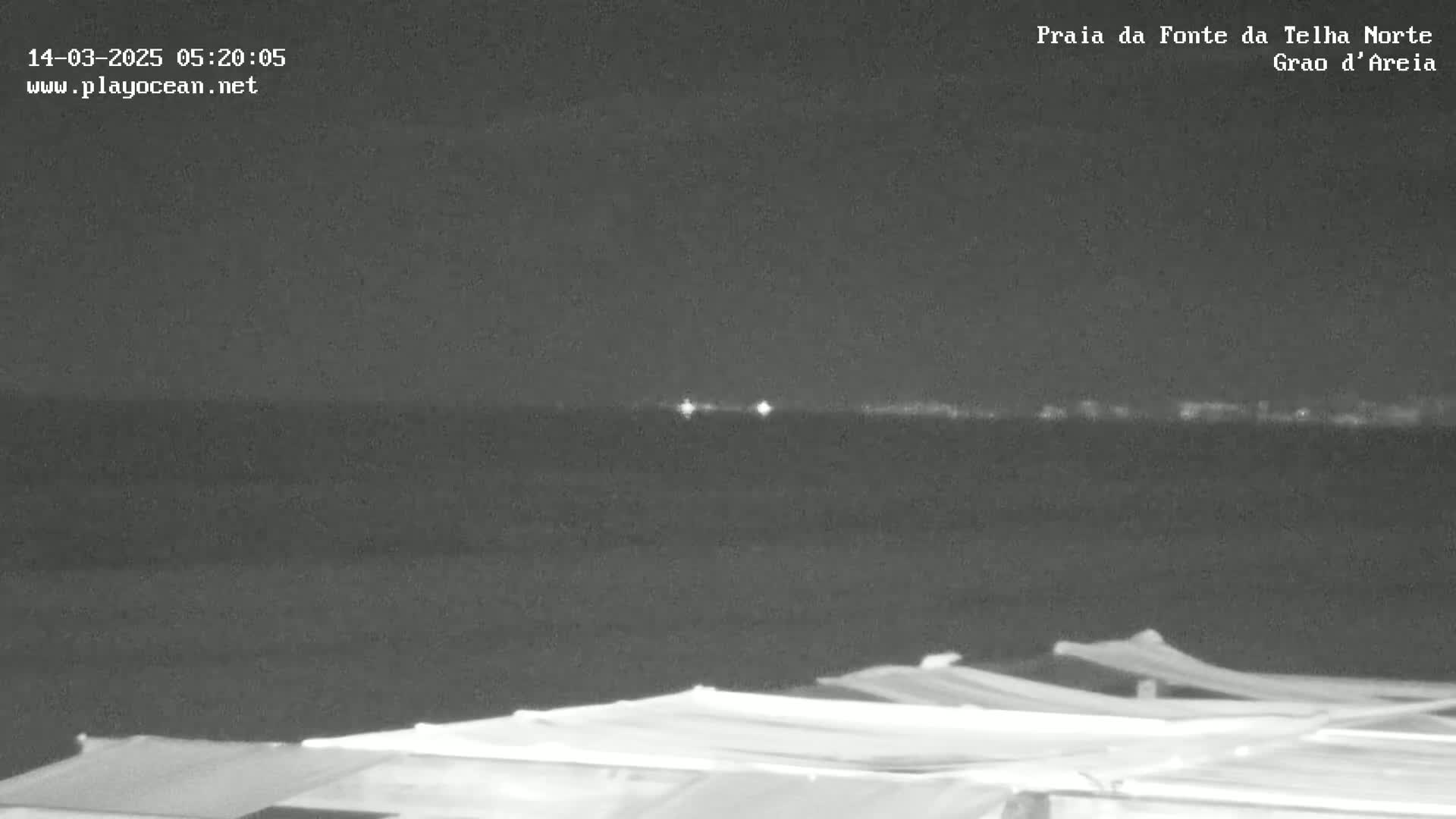 A nighttime view of a dark body of water with a distant, faintly lit shoreline, and a foreground of what appears to be a covered structure.
