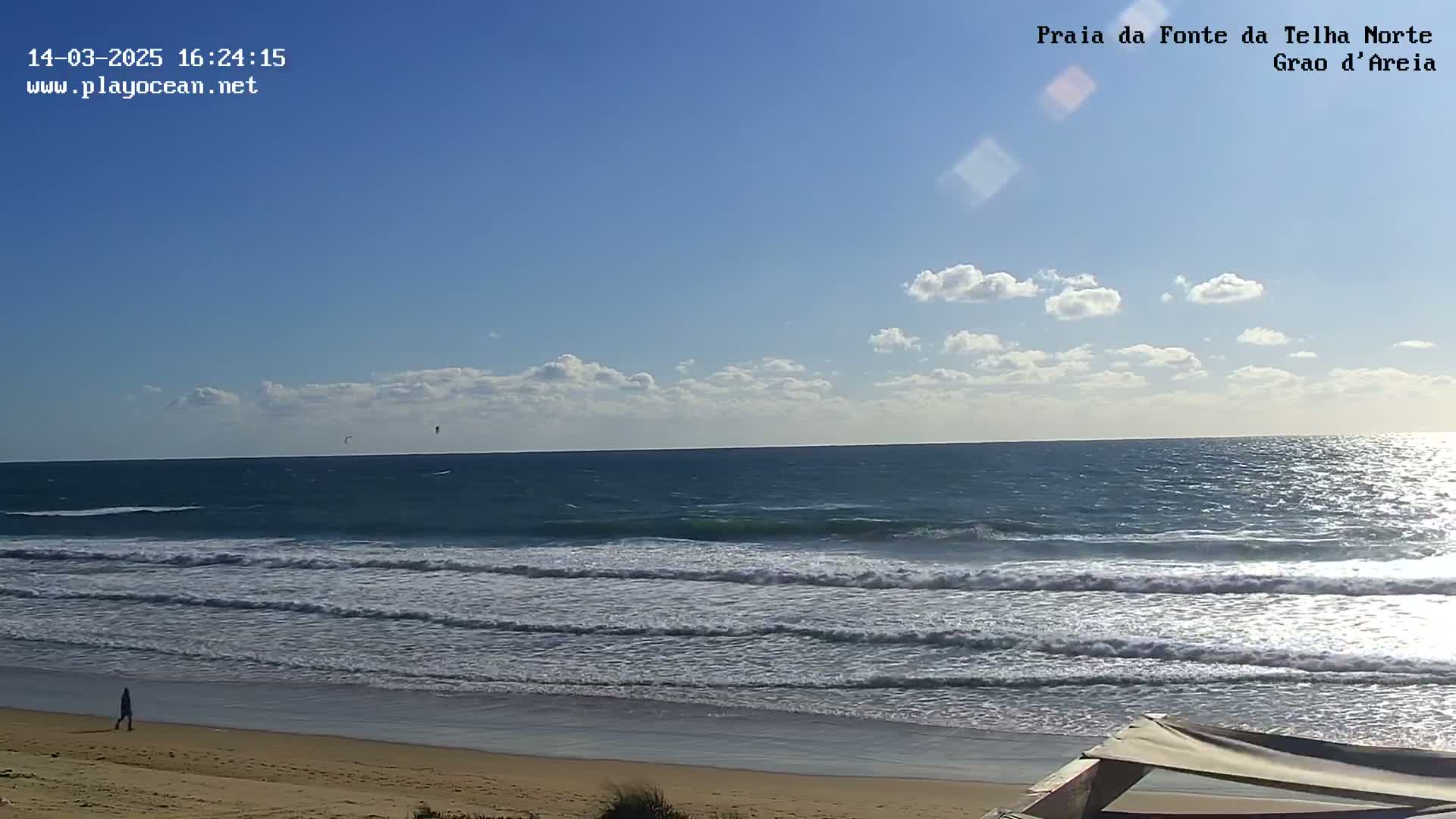 A person walks along a sandy beach next to a wavy ocean under a mostly sunny sky with a few scattered clouds.