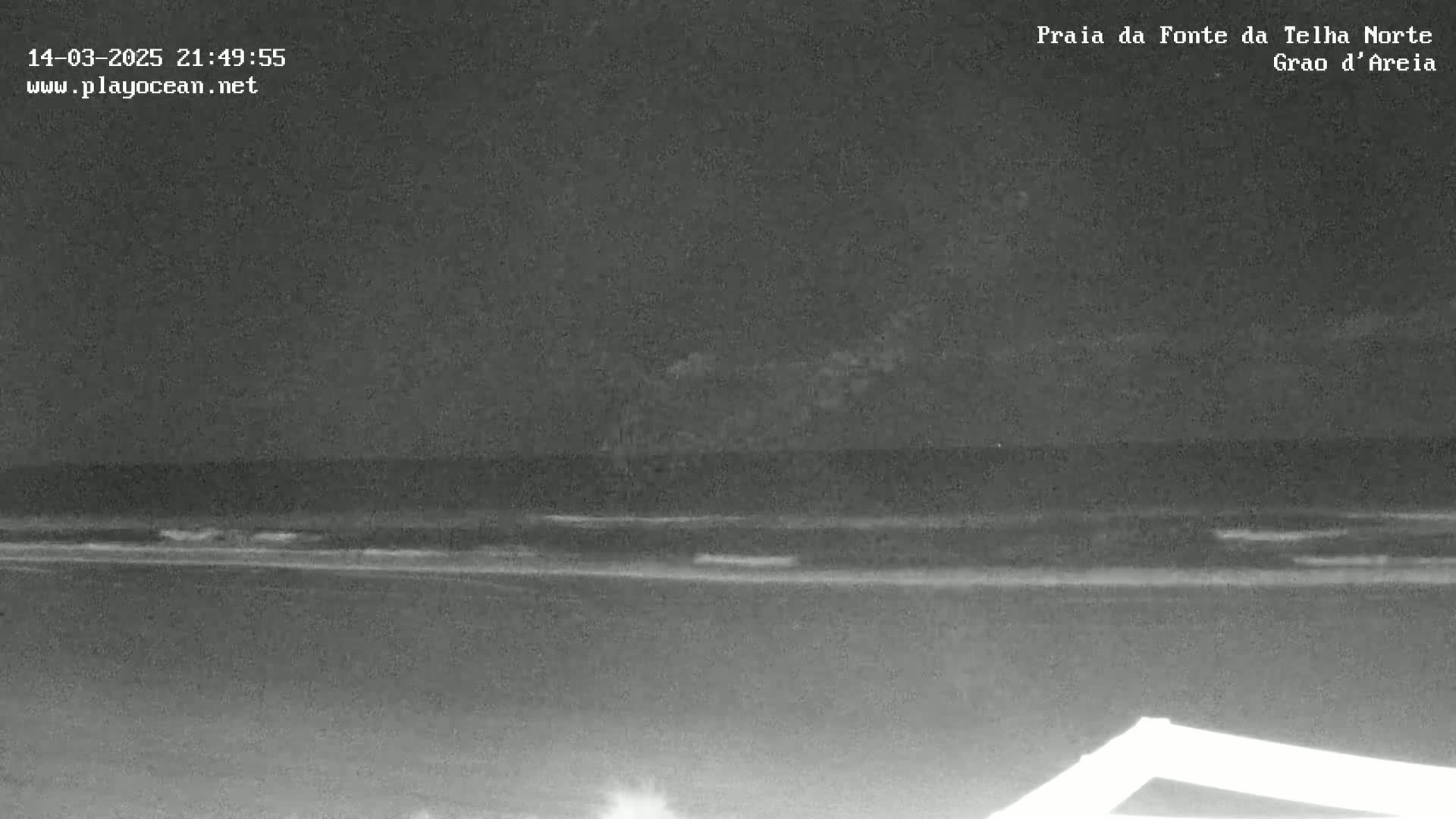 A nighttime, low-light image shows a dark ocean with small waves gently lapping a beach under a cloudy sky.