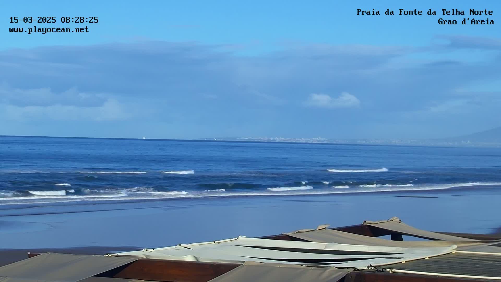 A sandy beach with a calm ocean and a mostly clear sky is viewed from behind a partially covered outdoor structure.