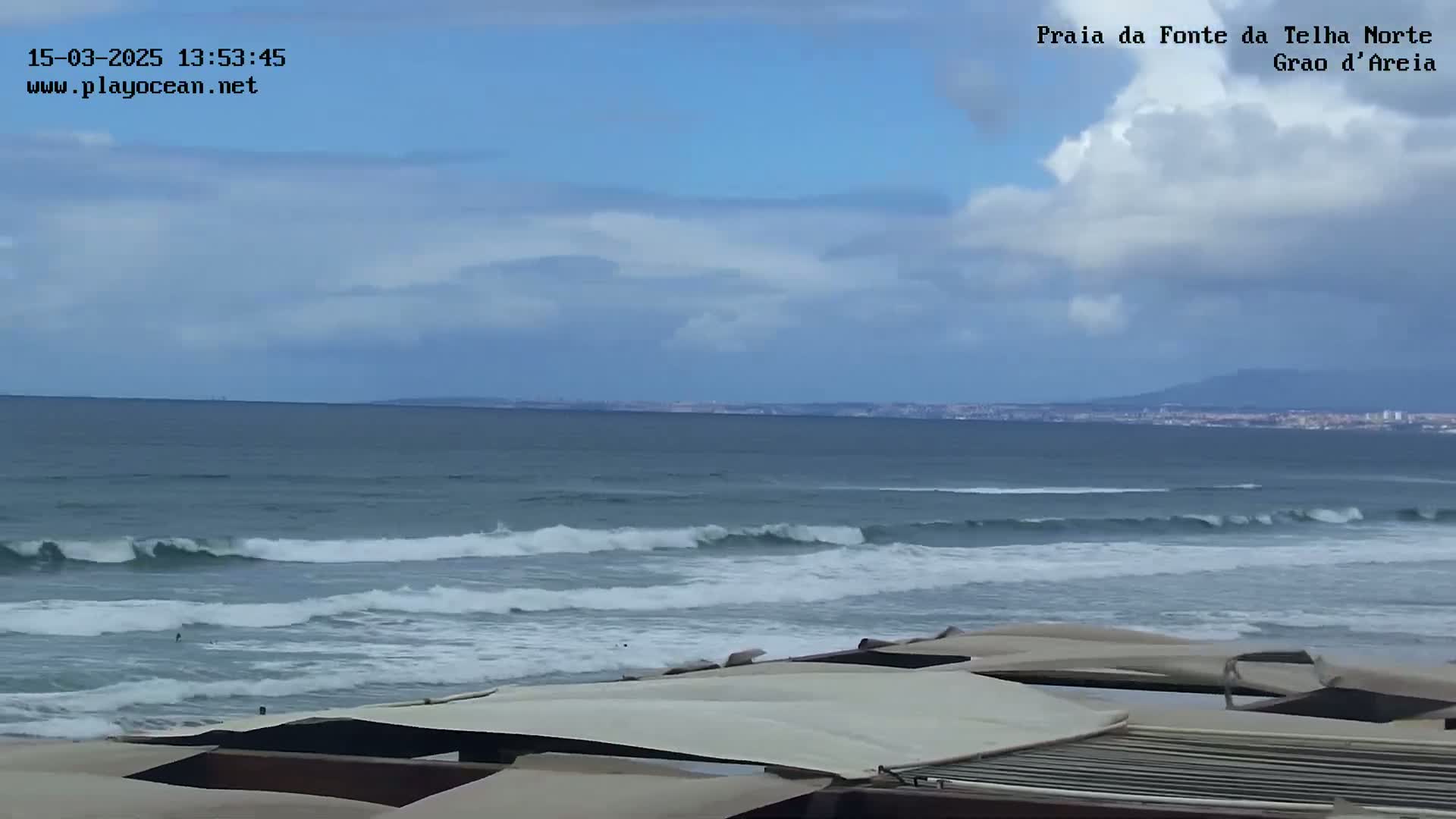 An ocean beach with gently rolling waves under a partly cloudy sky is viewed from behind a row of beach canopies.