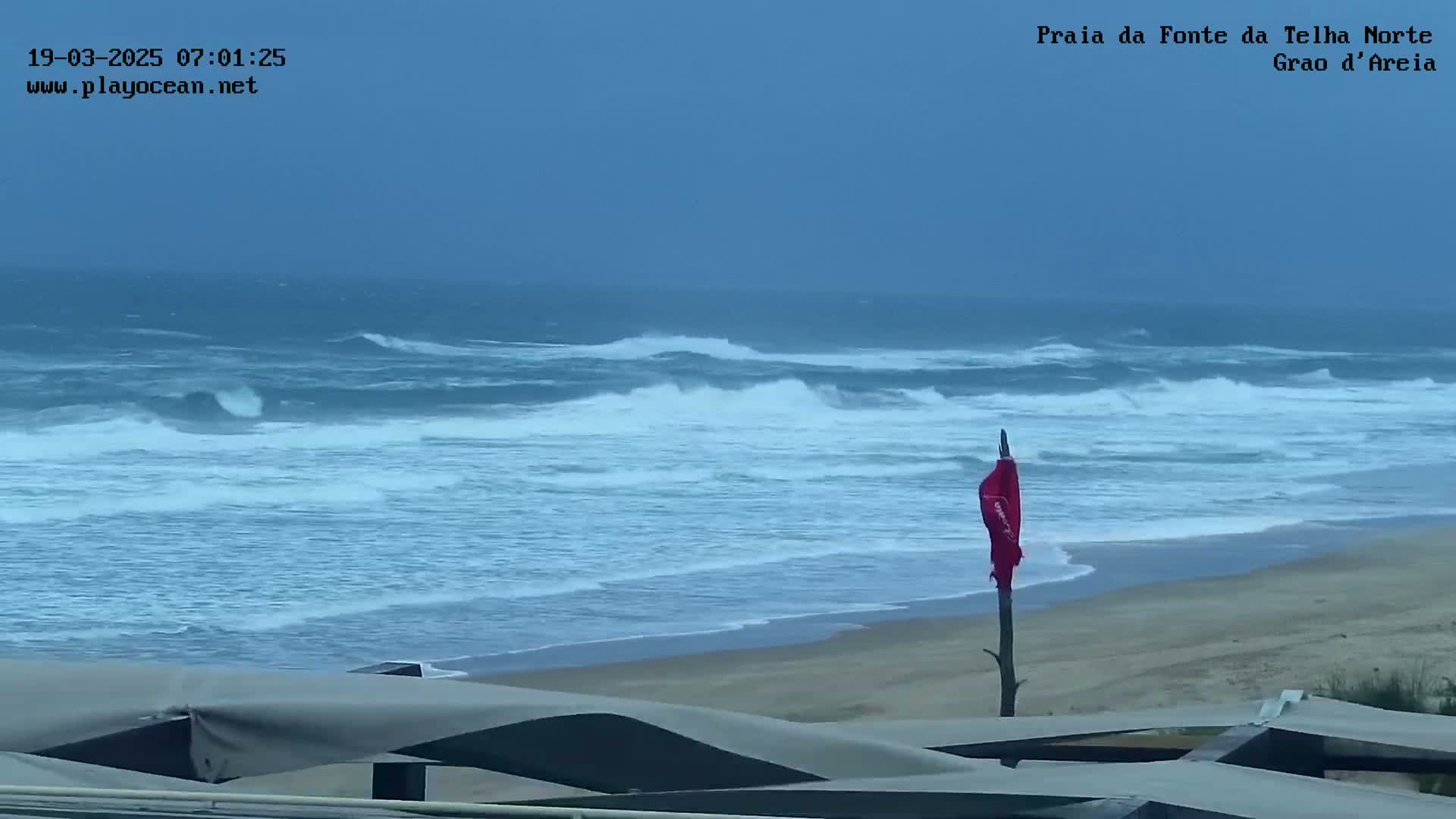 A mostly cloudy day shows a sandy beach with moderate waves and a red flag on a post in the foreground.