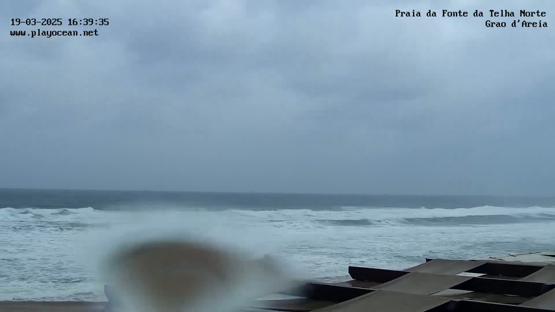 A beach scene shows choppy ocean waves under an overcast sky, with some dark-colored structures in the foreground.