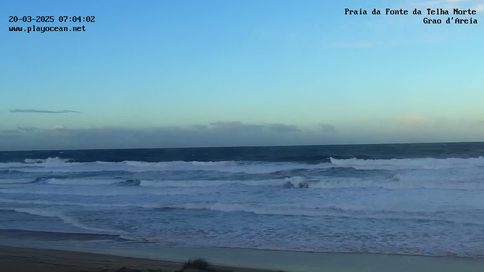 A wide, sandy beach is shown with moderately sized waves rolling into shore under a clear, light-blue sky.