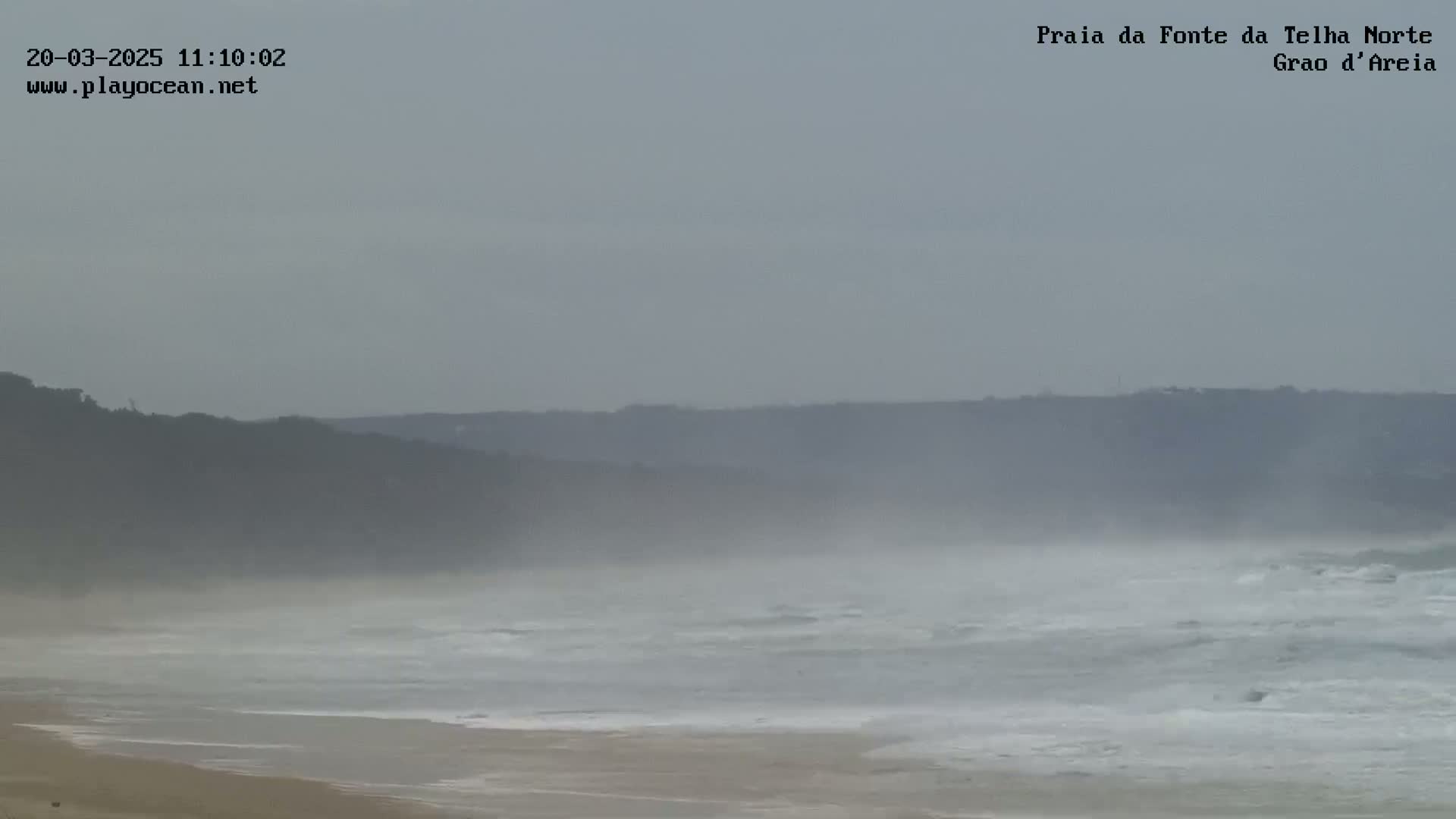 A hazy, overcast day shows a beach with moderate waves crashing on the shore, backed by a distant, indistinct cliff line.
