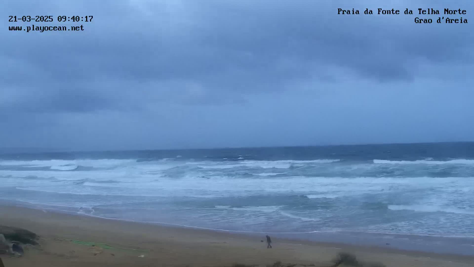 A person walks a dog on a sandy beach under an overcast sky as moderately sized waves crash on the shore.