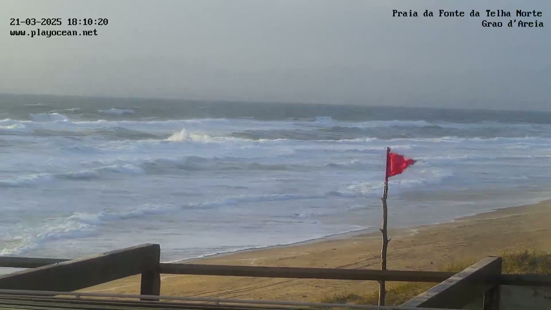 A mostly cloudy day shows a sandy beach with choppy waves and a red flag on a stick indicating potentially hazardous conditions.