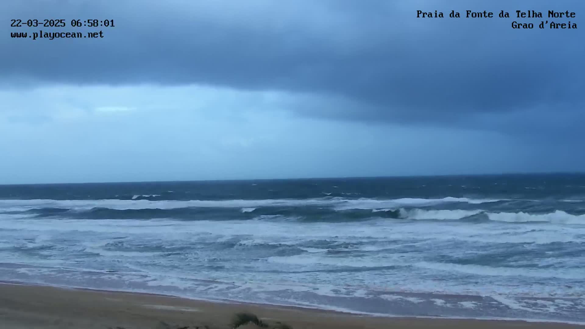 A sandy beach meets a dark, choppy ocean under a stormy, overcast sky.