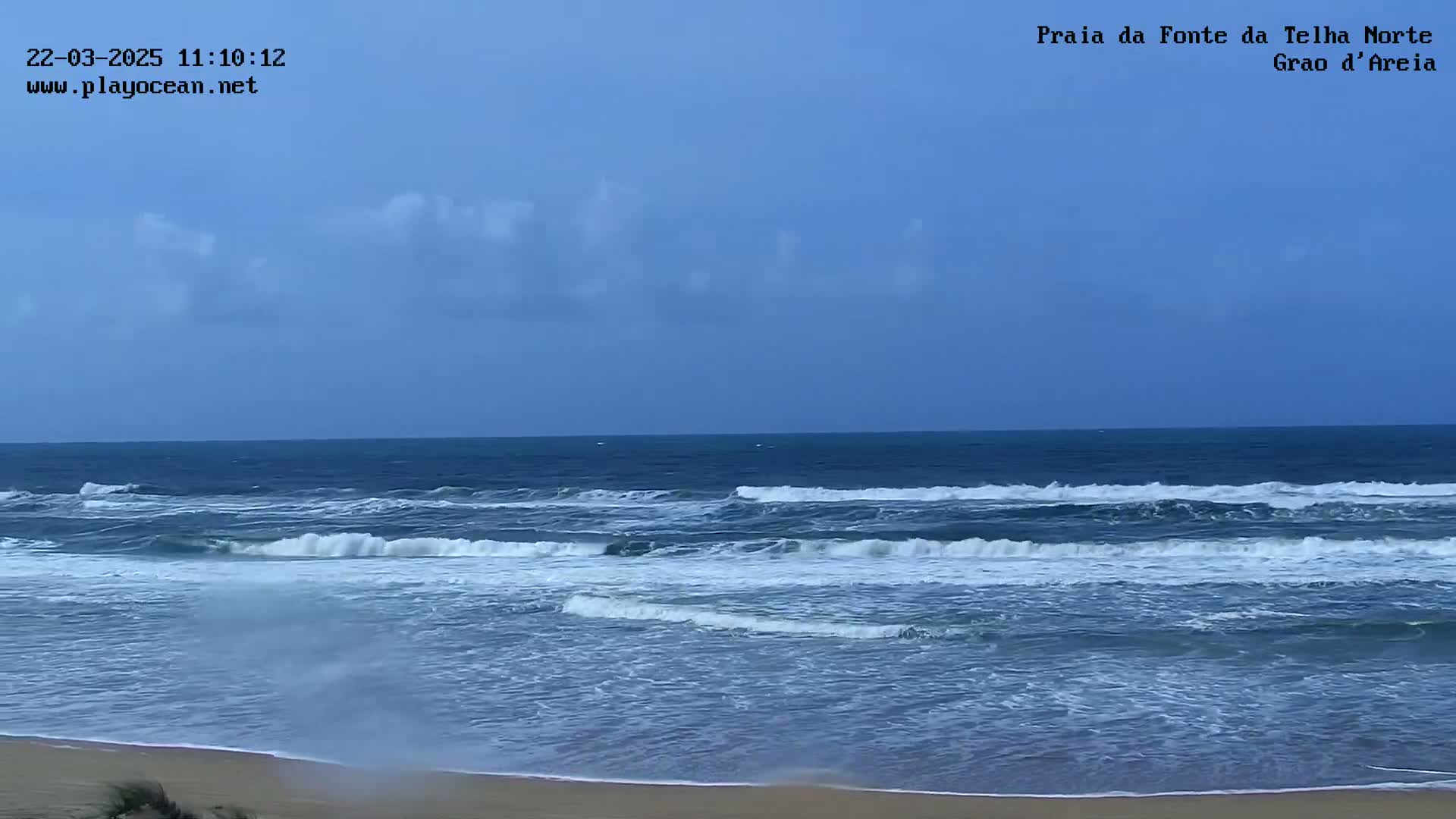 A partly cloudy sky overlooks a beach with moderate waves breaking onto a sandy shore.