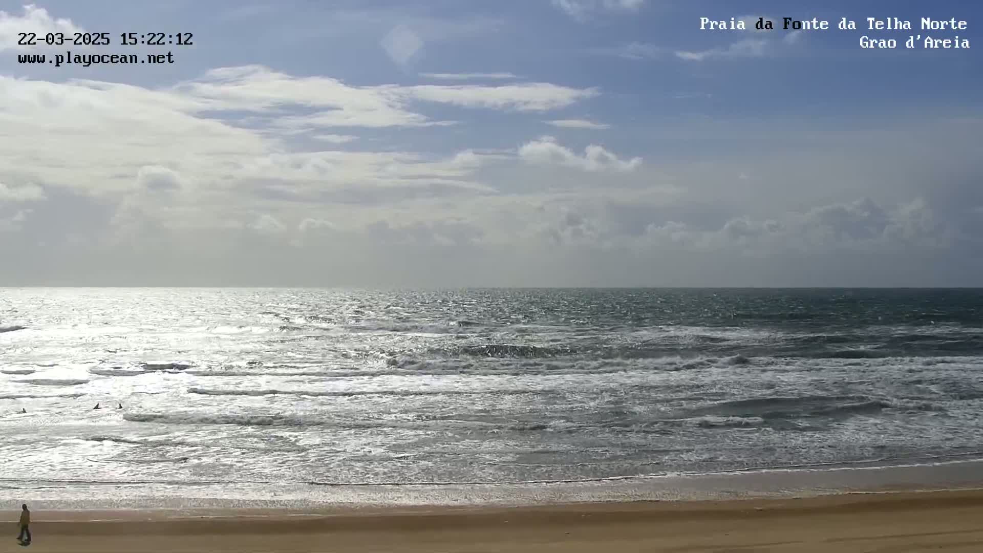 A person walks on a sandy beach under a partly cloudy sky, overlooking a moderately wavy ocean.