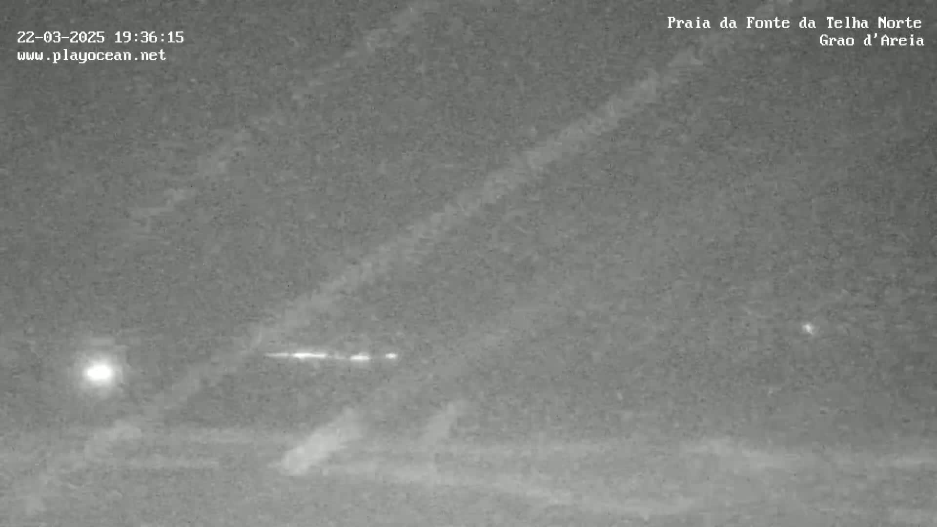 A nighttime, low-light image shows a dark, possibly sandy beach with a faint light source in the distance and a bright streak of light across the sky.