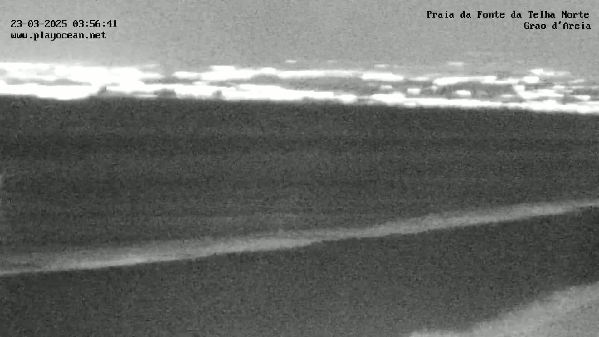 A low-light, grainy image shows a dark beach and ocean under a mostly cloudy sky.
