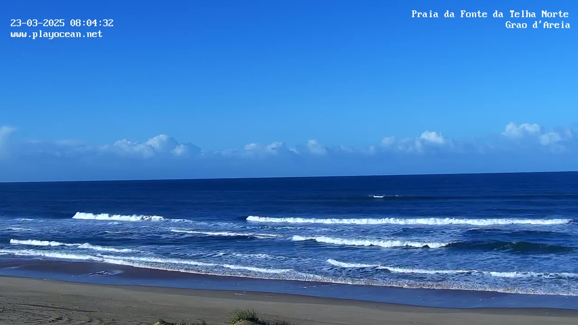 A sandy beach meets a calm, blue ocean under a clear sky with a few scattered clouds.