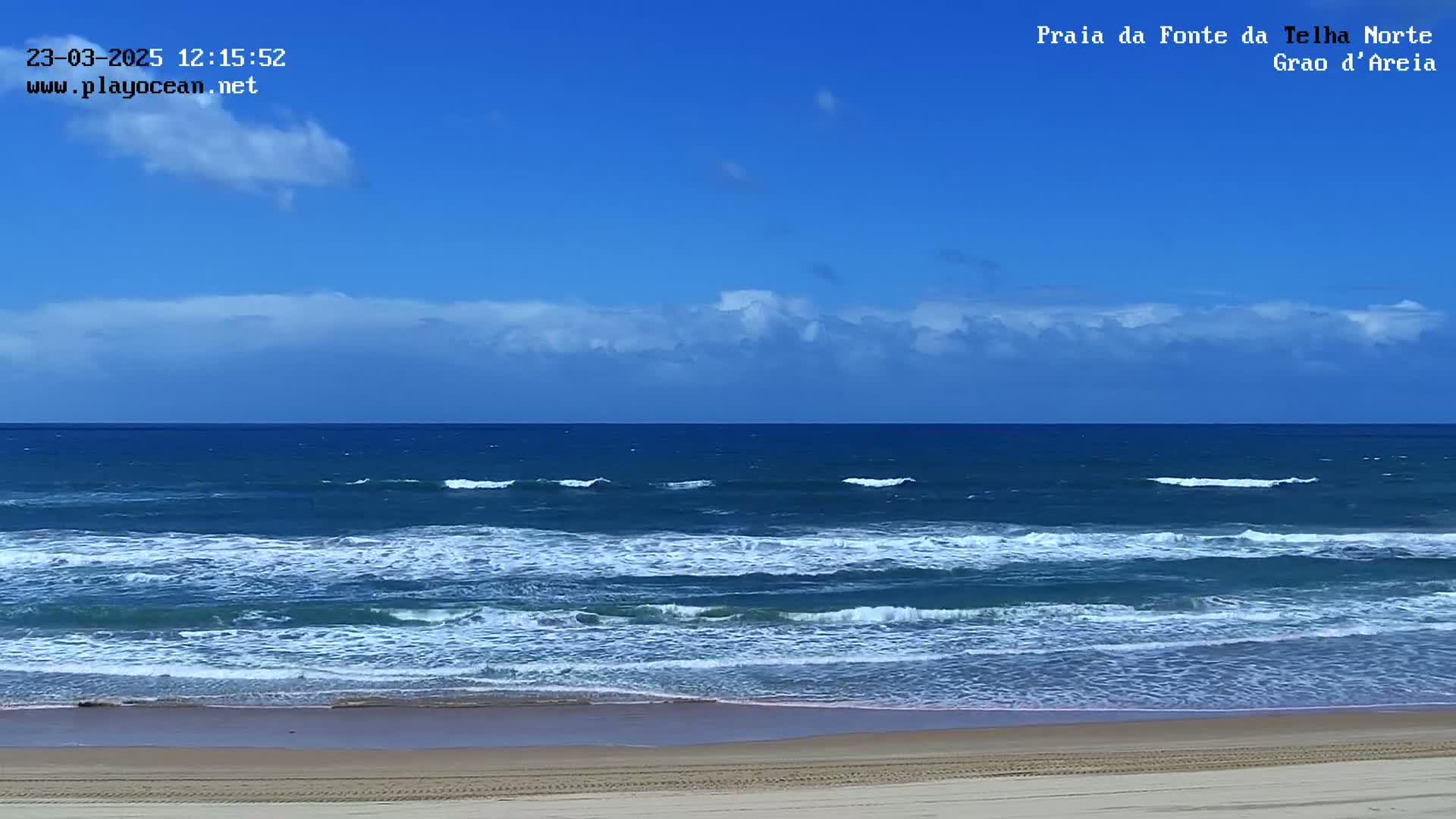A sandy beach meets a blue ocean with moderate waves under a mostly sunny sky with some scattered clouds.