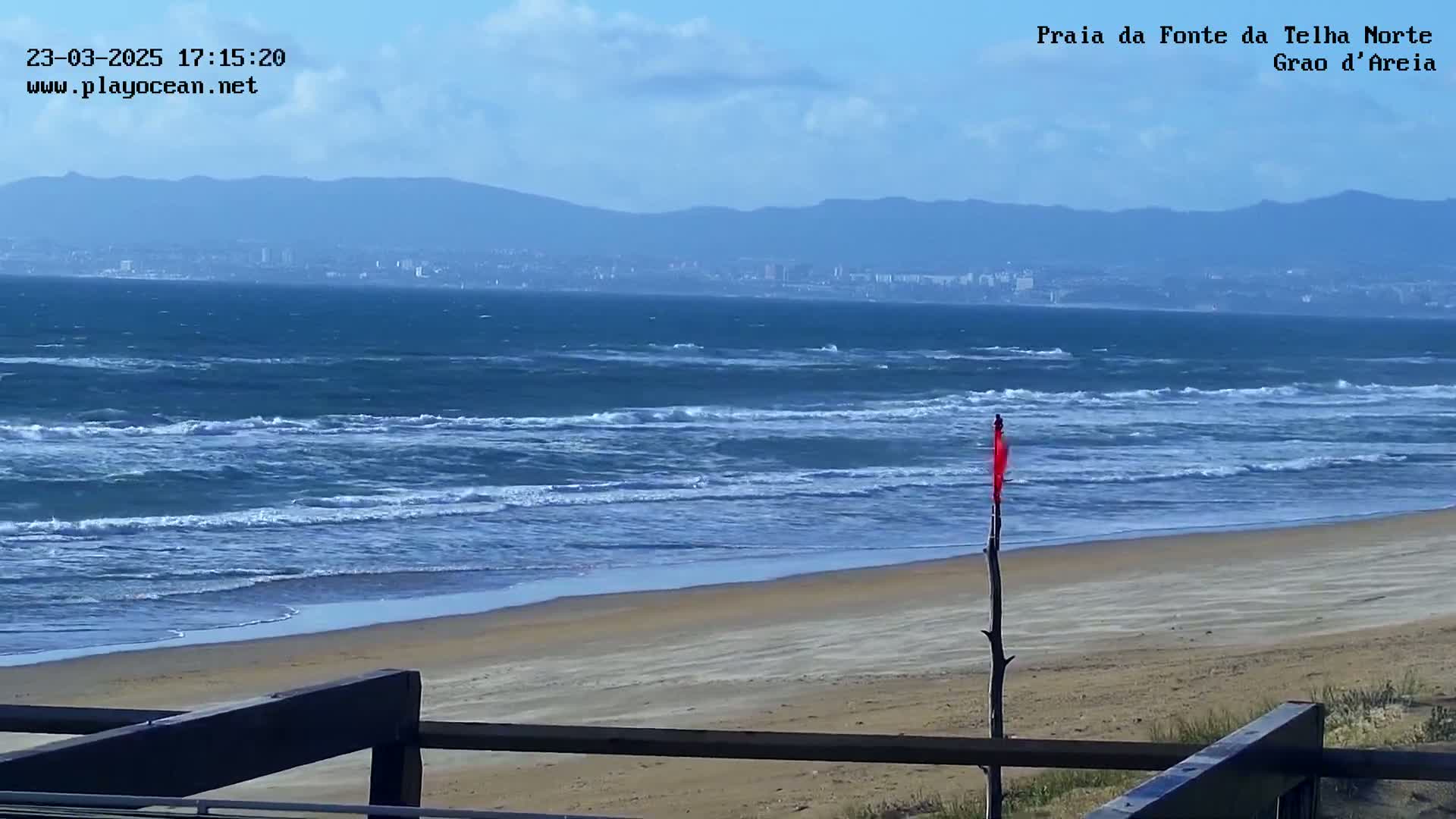 A partly cloudy day shows a sandy beach with choppy blue ocean waves, a distant city, and mountains under a mostly clear sky.