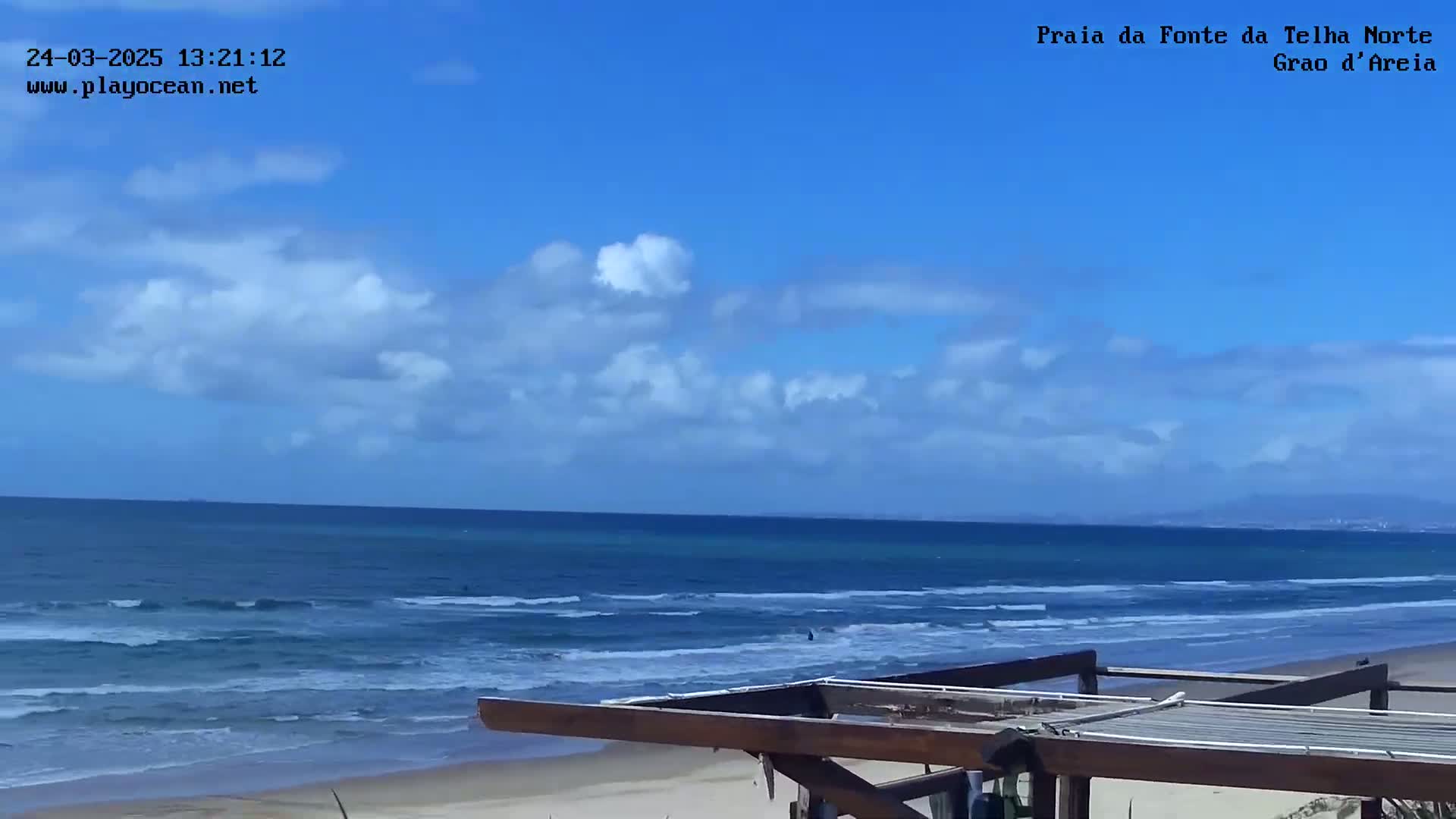 A partly cloudy blue sky overlooks a tranquil ocean with small waves lapping a sandy beach, partially obscured by a wooden structure in the foreground.