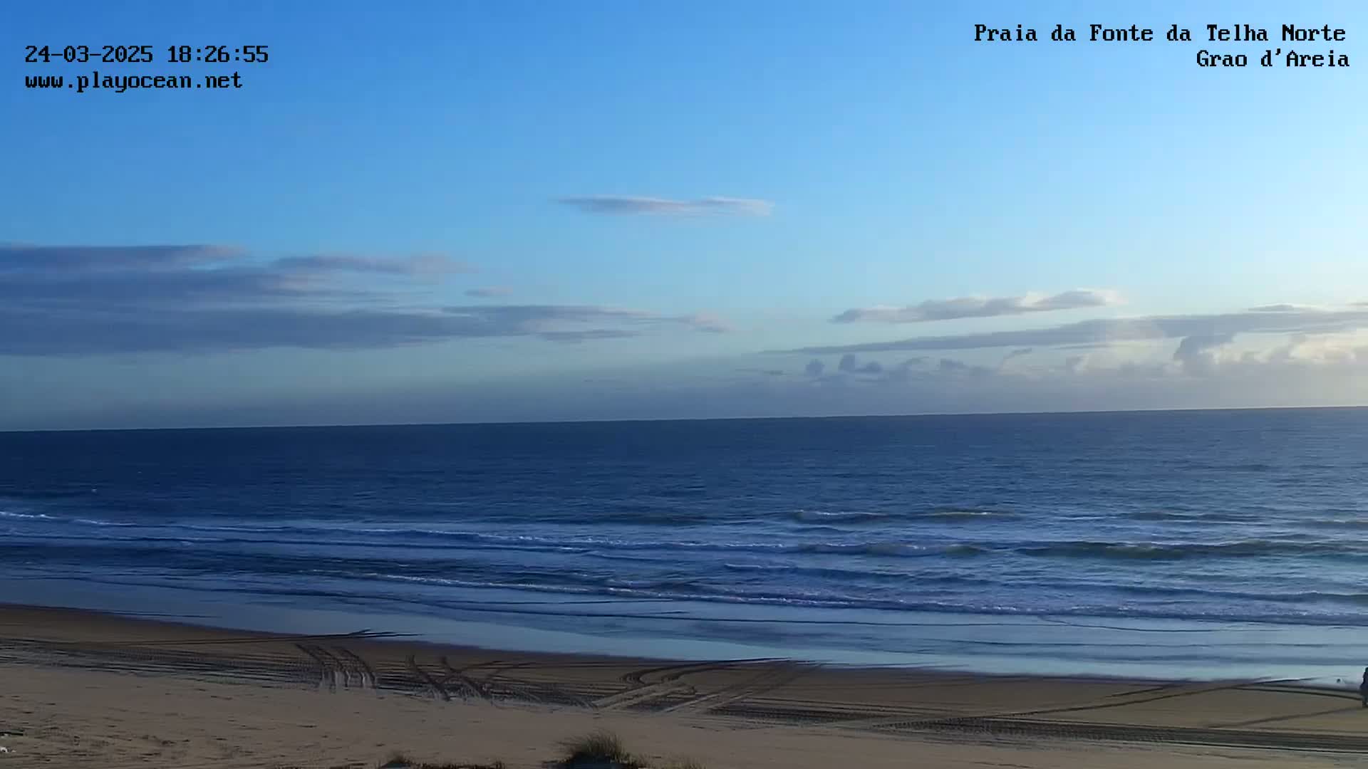 A mostly clear blue sky over a calm ocean meeting a sandy beach with tire tracks.