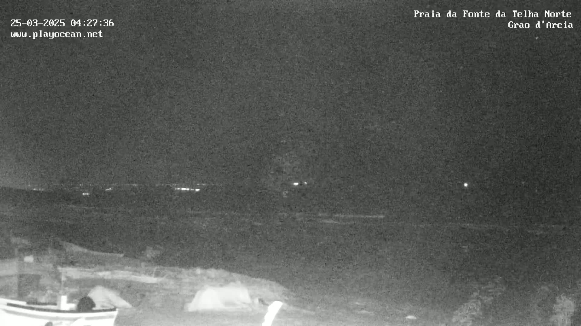 A nighttime, low-light image shows a dark, possibly sandy shore with a boat and some objects that appear to be tents in the foreground, and a dark body of water extending to a distant, faintly lit horizon under a clear night sky.