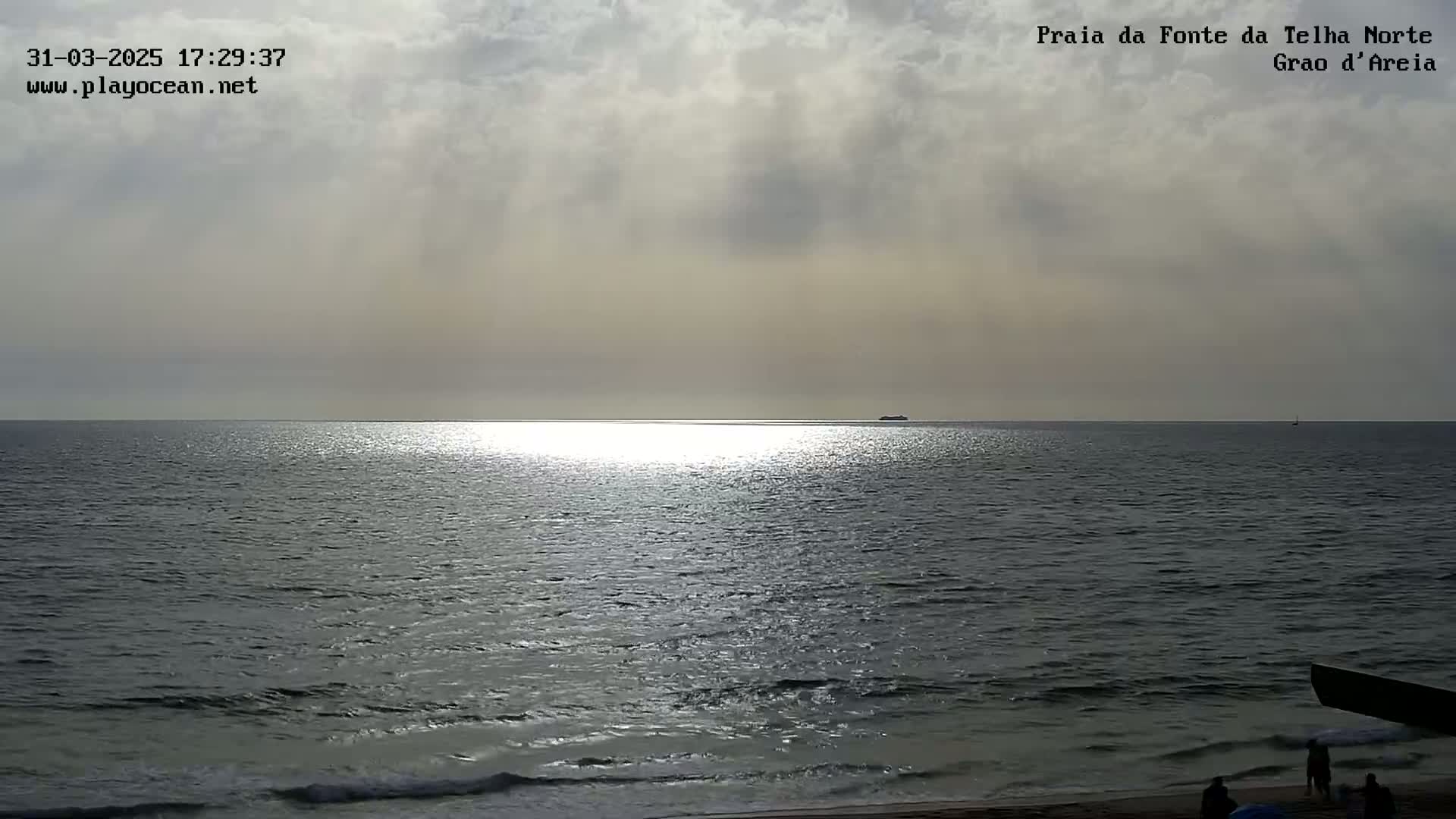 A mostly cloudy day shows a calm ocean reflecting sunlight on the water near a sandy beach where several people are walking.
