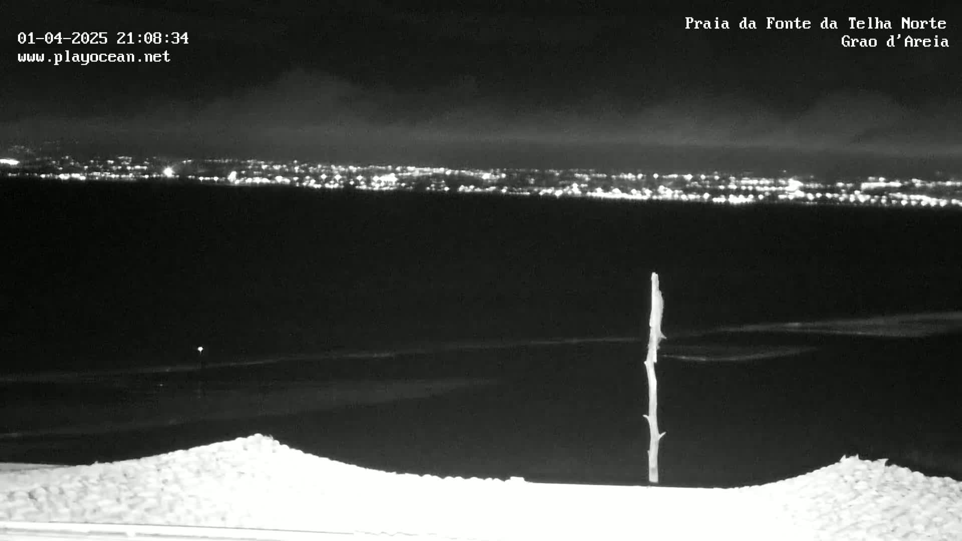 A nighttime view of a distant city skyline illuminated with lights, seen from a dark, possibly coastal area with a single, thin, upright post in the foreground.