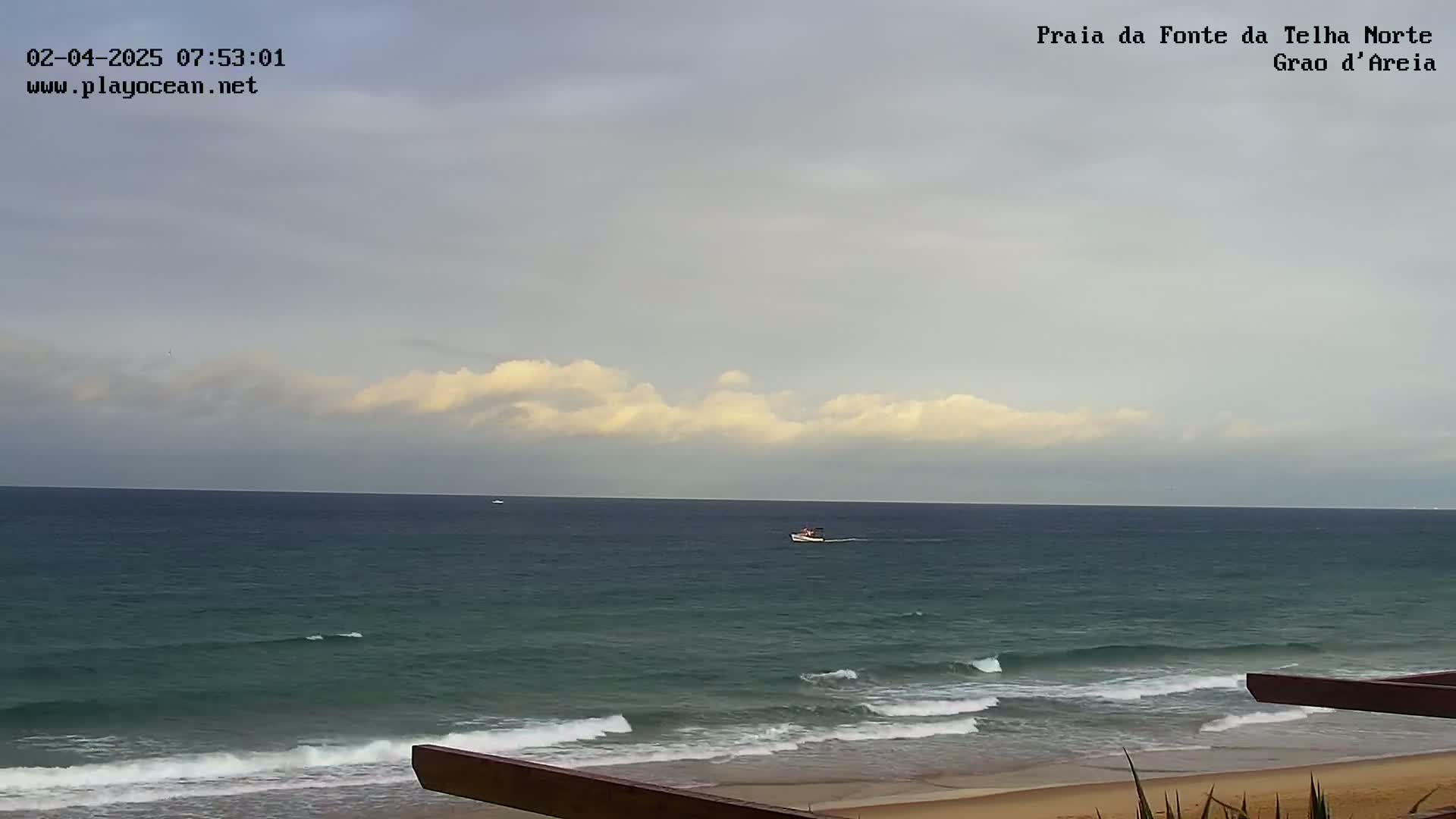 A mostly cloudy sky overlooks a calm ocean with small waves lapping a sandy beach.
