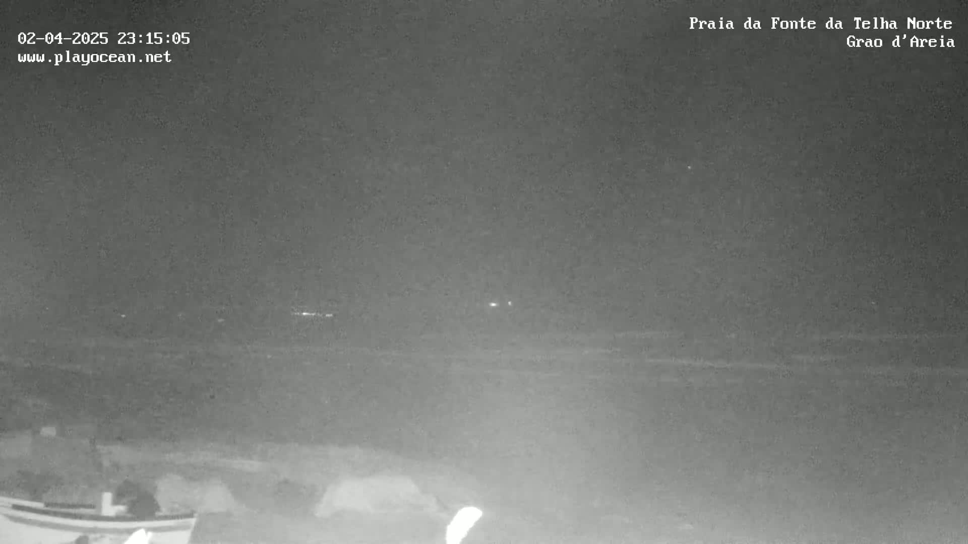 A nighttime, low-light view of a dark ocean shore with faint lights visible in the distance.