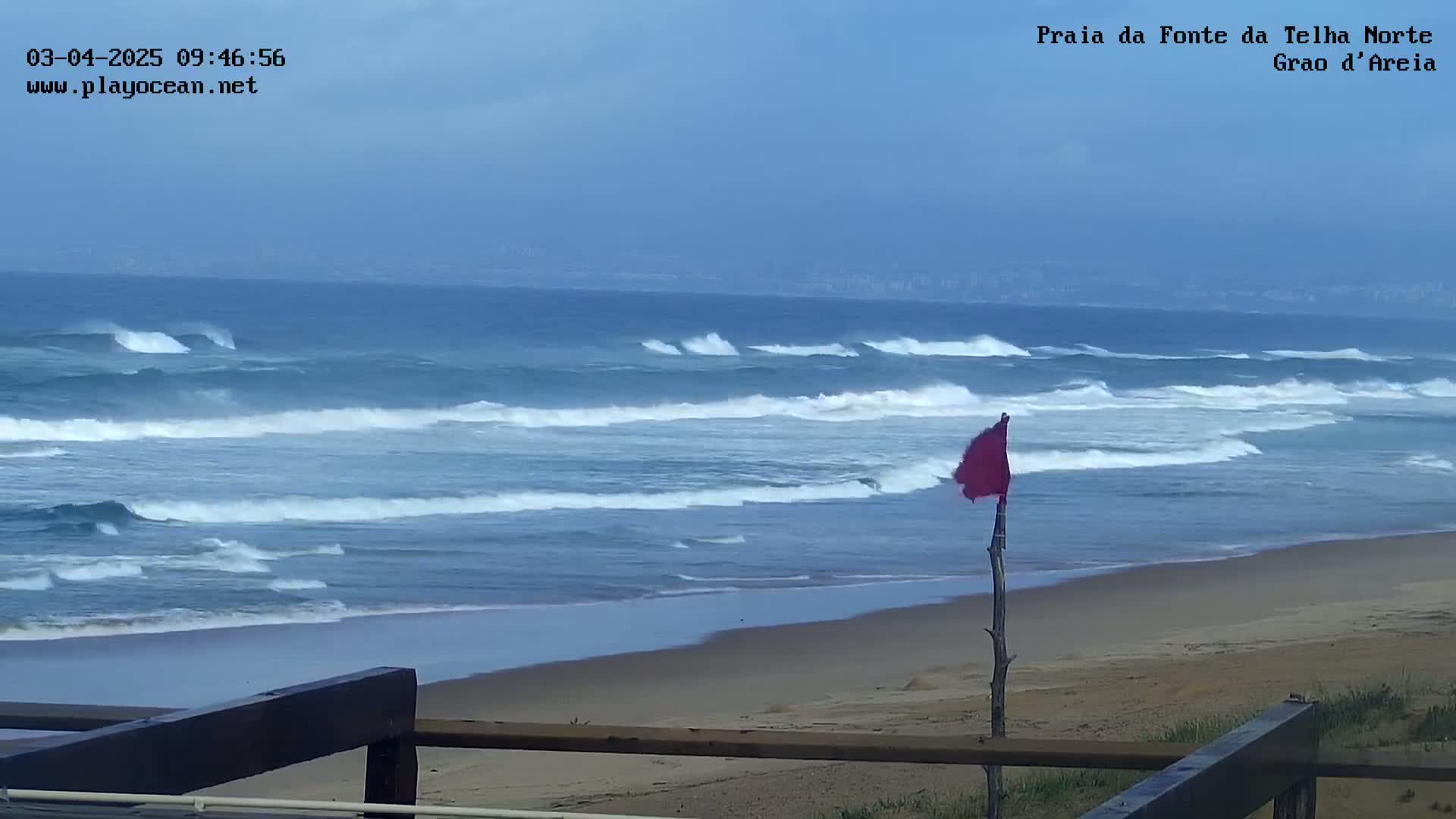 A sandy beach with moderate waves under an overcast sky features a red flag on a wooden post in the foreground.