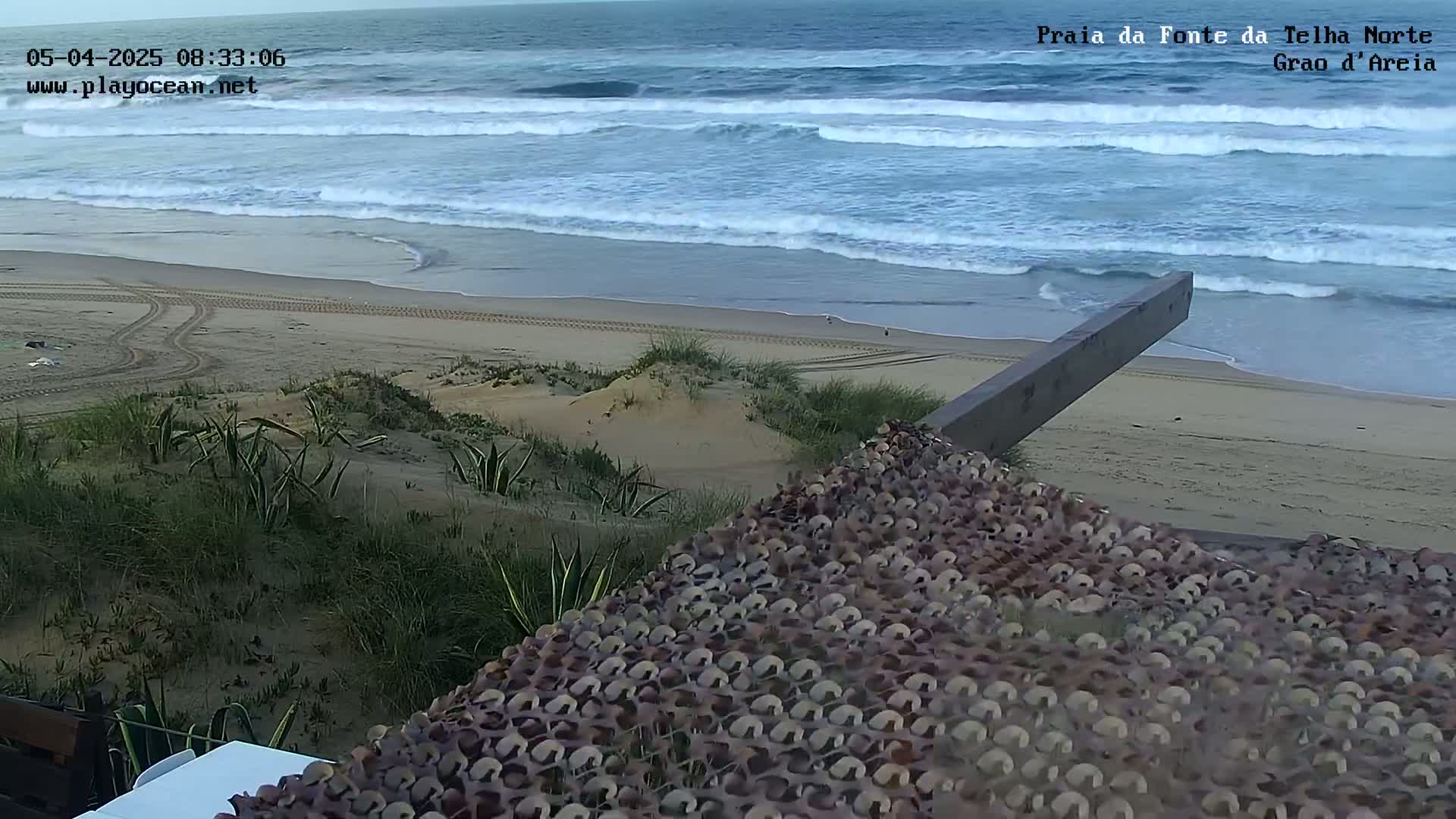 A sandy beach with gentle waves under a cloudy sky is viewed from a dune overlooking a camouflaged structure.