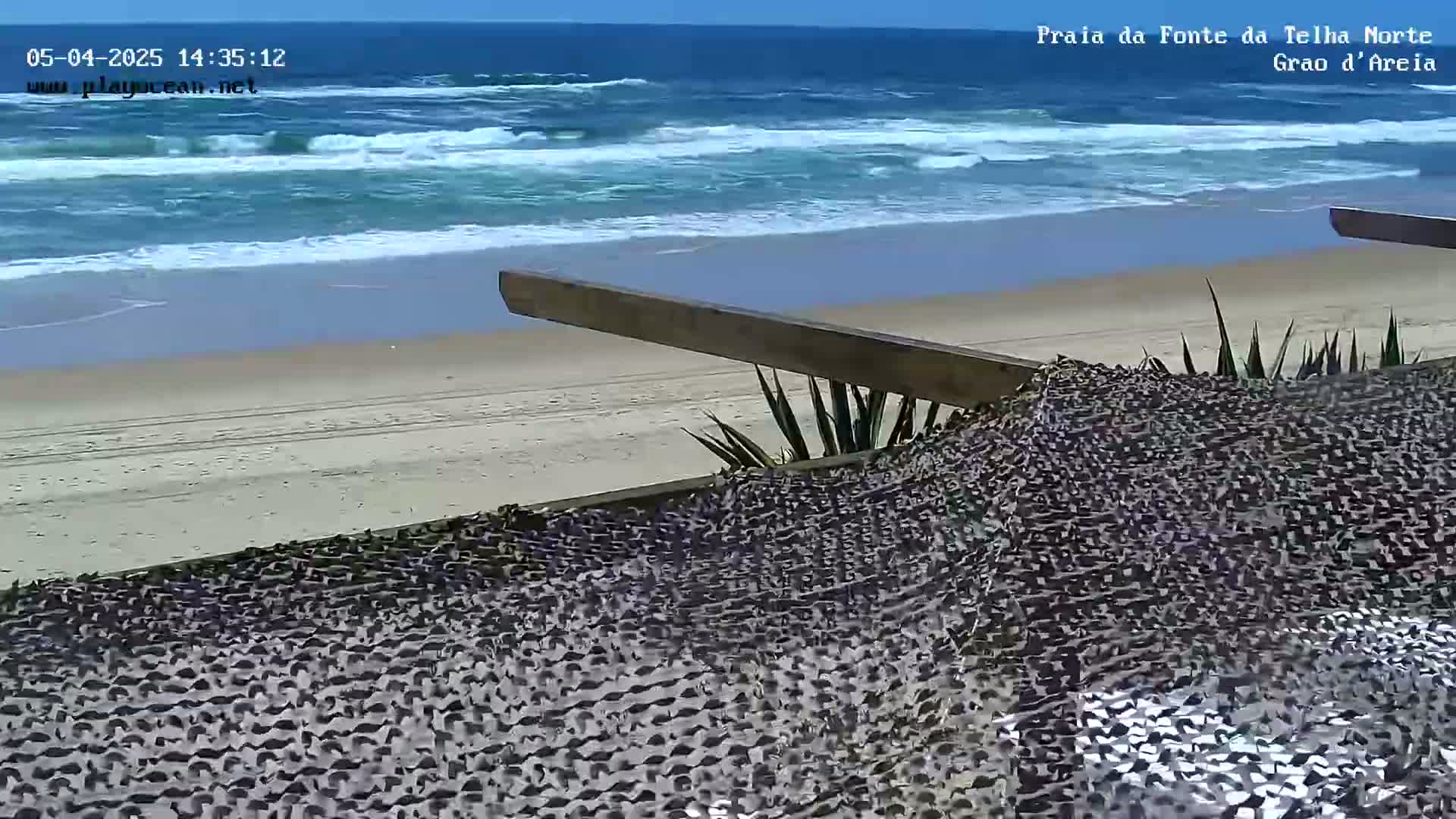 A sandy beach with moderate waves under a clear sky is viewed from behind a textured fabric covering a wooden structure.