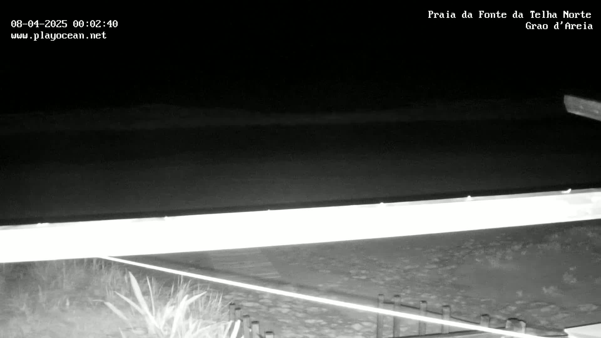 A nighttime, low-light view of a beach with a dark ocean and a long, bright structure near the shore.