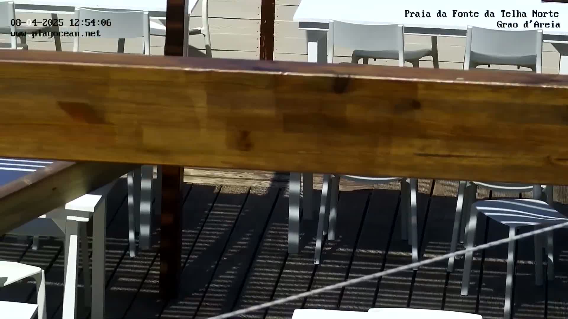 A wooden beam partially obscures the view of a sunny outdoor patio with white tables and chairs on a dark-colored deck.