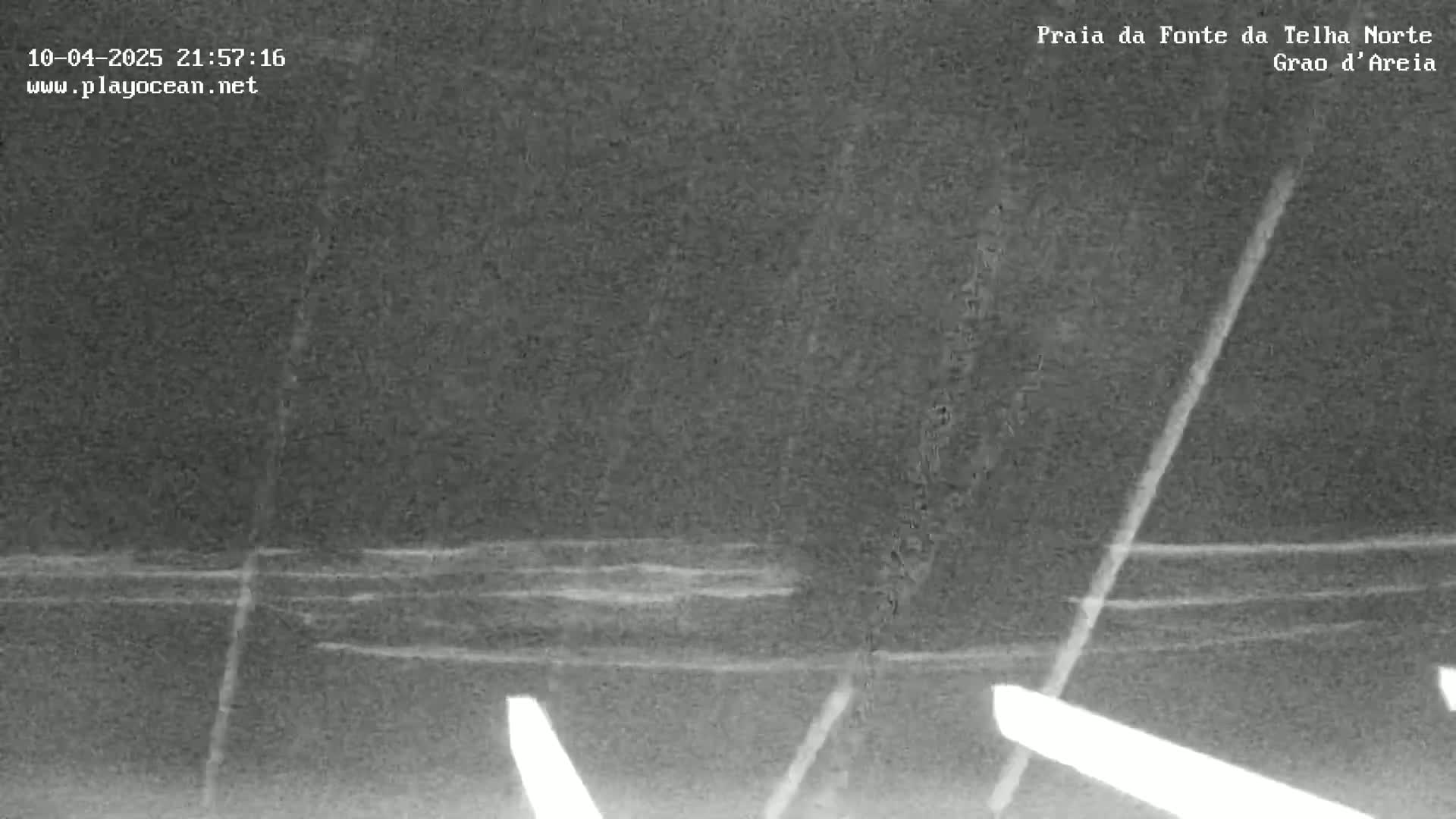 A nighttime, low-light image shows dark water with faint, parallel lines suggesting waves, and bright lines on the land indicating lights or reflections.