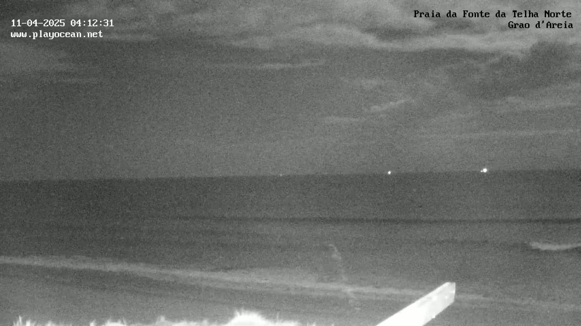 A nighttime, low-light view shows a dark ocean meeting a dark sandy beach under a cloudy sky.